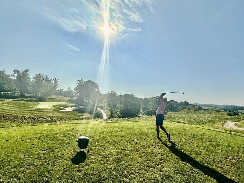 A golfer teeing off with the sun flaring behind the trees