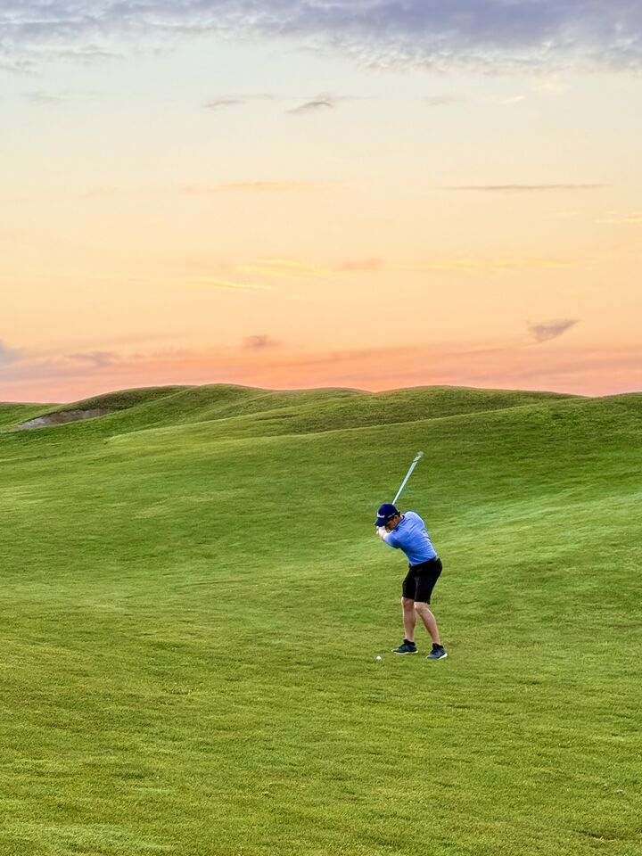 A golfer finishing a swing on an open fairway at sunset